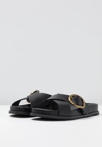 Černé kožené sandály s širokými křížovými popruhy a velkým zlatým kováním jako akcentem. Plochá podrážka se hladkým povrchem a minimalistickým designem.