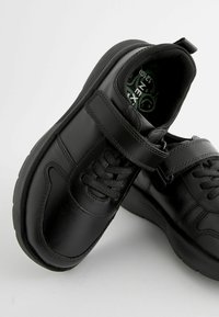Chaussures en cuir noir avec une texture lisse, dotées d'une bride en Velcro et de lacets traditionnels, semelle en caoutchouc texturée et une semelle intérieure à motif vert.