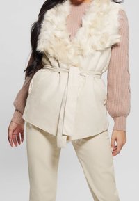 Donna che indossa un gilet con colletto in pelliccia sintetica beige con cintura sopra un maglione a maniche lunghe rosa chiaro a coste e pantaloni crema.