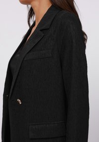 Černý blazer s texturovanou tkaninou, strukturovaným klopem, jednou náprsní kapsou a zlatými detaily na přední straně.
