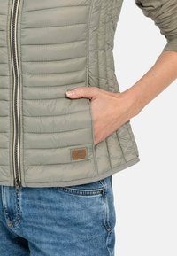 Jasnoszara, pikowana kamizelka z zamkiem błyskawicznym z przodu i bocznymi kieszeniami. Posiada fakturowany materiał oraz brązową łatkę z logo na talii.