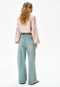Kobieta o długich falujących włosach, ubrana w beżową bluzkę z bufkimi rękawami i szerokie jasnoniebieskie dżinsy, stojąca tyłem na białym tle.