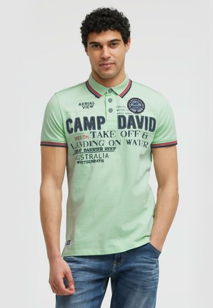 Mann mit lockigem Haar, der ein hellgrünes Poloshirt mit dem Text "Camp David" und blaue Jeans trägt, steht mit einer Hand in der Tasche vor einfarbigem Hintergrund.