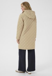 Manteau matelassé beige avec capuche, présentant un motif en losanges et des manches longues. Associé à un pantalon large sombre et des baskets blanches.