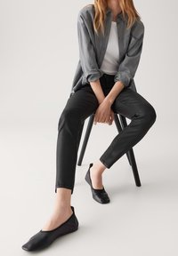 Personne assise sur une chaise noire portant une chemise grise boutonnée, un top blanc, un pantalon noir et des chaussures plates noires sur un fond blanc uni.