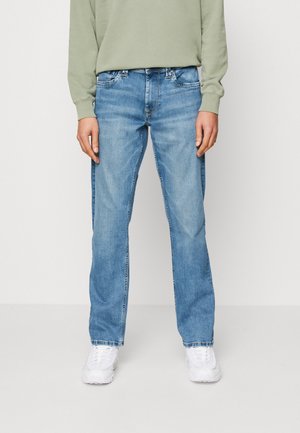 Persona che indossa jeans azzurri chiari a gamba dritta, sneakers bianche e una felpa verde chiaro, in piedi davanti a uno sfondo bianco.
