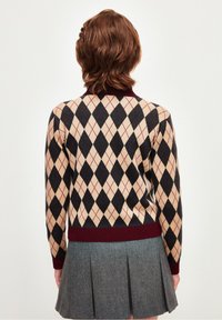 Maglione lavorato a maglia con un motivo a diamante in beige, nero e bordeaux. Collo e polsini a costine, indossato con una gonna plissettata grigia.