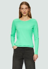 Dolgi rokavi, majica s krojem okroglega vratu v svetlem meta zelenem bombažu. Gladka tekstura, prilegajoča zasnova, v kombinaciji s temnimi kavbojkami, roki v žepih.