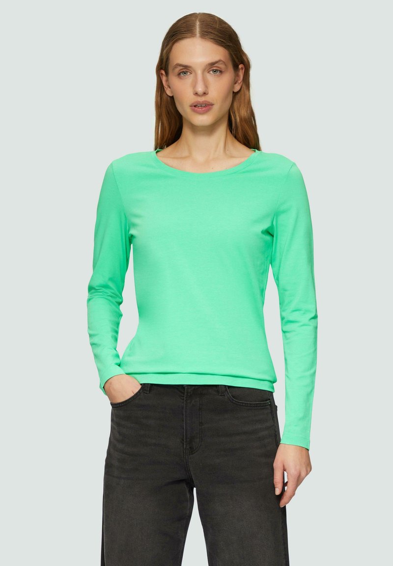 Dolgi rokavi, majica s krojem okroglega vratu v svetlem meta zelenem bombažu. Gladka tekstura, prilegajoča zasnova, v kombinaciji s temnimi kavbojkami, roki v žepih.