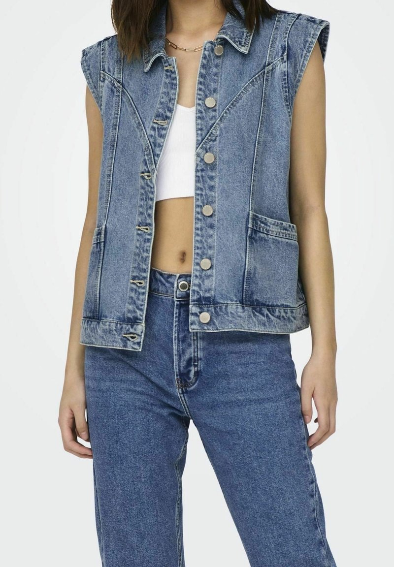 Džínová vesta bez rukávů, světle modré barvy, s předními kapsami a stříbrnými vrchními knoflíky. Nositelná přes bílý crop top.