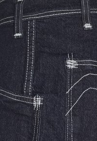 Tumma denimkangas, jossa on valkoiset ompeleet, etutasku ja huomaamattomat kuluneisuuden yksityiskohdat pinnassa. Lähellä oleva kuva kankaasta.