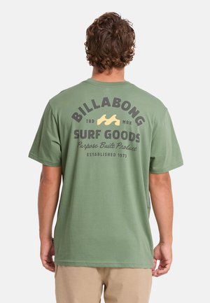 Mann trägt grünes Billabong Surf Goods T-Shirt mit Logo und Schriftzug auf dem Rücken, beige Shorts, von hinten auf einfarbigem Hintergrund gesehen.