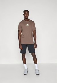 Ruda medvilninė marškinėliai su auksiniu apvaliu Nike logotipu, derinama su tamsiomis trumpomis kelnėmis. Modelis dėvi pilkas sportines batus su baltais akcentais.