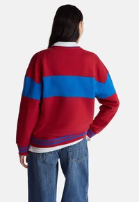 Czerwona i niebieska bluza z blokami kolorów o luźnym kroju, z prążkowanymi mankietami i pasem, oraz białym kołnierzem, widoczna od tyłu.