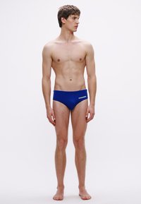 Blaue Badehose aus dehnbarem Stoff, mit einem niedrig geschnittenen Design und einem weißen Logo auf der linken Seite. Glatte Textur, minimaler Stoffeinsatz.