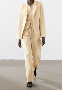 Jasnobeżowy garnitur składający się z dopasowanego blezera i spodni o szerokich nogawkach z teksturowanym wykończeniem. Uzupełniony o kamizelkę na guziki. Widoczne czarne sandały.