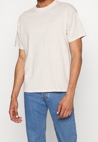 Jasno beżowa bawełniana koszulka z okrągłym dekoltem i krótkimi rękawami, z subtelnym logo na klatce piersiowej, w połączeniu z niebieskimi dżinsami.