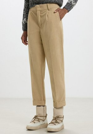 Pantalon en velours côtelé beige avec une taille haute, un devant plissé et des ourlets retroussés, associé à des bottines beige. Texture lisse, coupe droite.