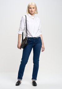 Camisa blanca de botones, jeans azules, bolso negro. Modelo lleva mocasines negros, cabello corto rubio, de pie contra un fondo gris claro.