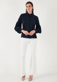 Blusa blu navy con bottoni frontali, colletto a punta e maniche a sbuffo. Abbinata a pantaloni bianchi a zampa d'elefante e tacchi argento texturizzati.