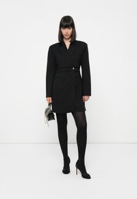 Sorte skreddersydd blazer-kjole med notched krage, enkelt knappelukking, sidelommer og lange ermer. Båret med sorte strømpebukser og hæler.