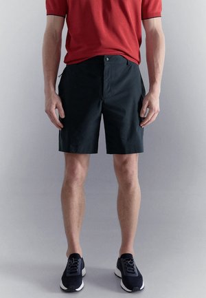 Homme portant un short noir longueur genoux, une chemise rouge à manches courtes et des baskets noires, debout devant un fond uni clair.