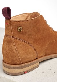 Botín de ante marrón hasta el tobillo con puntera redondeada, suela en un marrón claro contrastante, acento dorado del logo y una pestaña de tirador en burdeos en el talón.