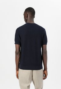 Mornarski pleten pulover s kratkimi rokavi, okroglim izrezom in rebrastimi manšetami. Teksturirana tkanina. Oblečen z svetlim hlačami.