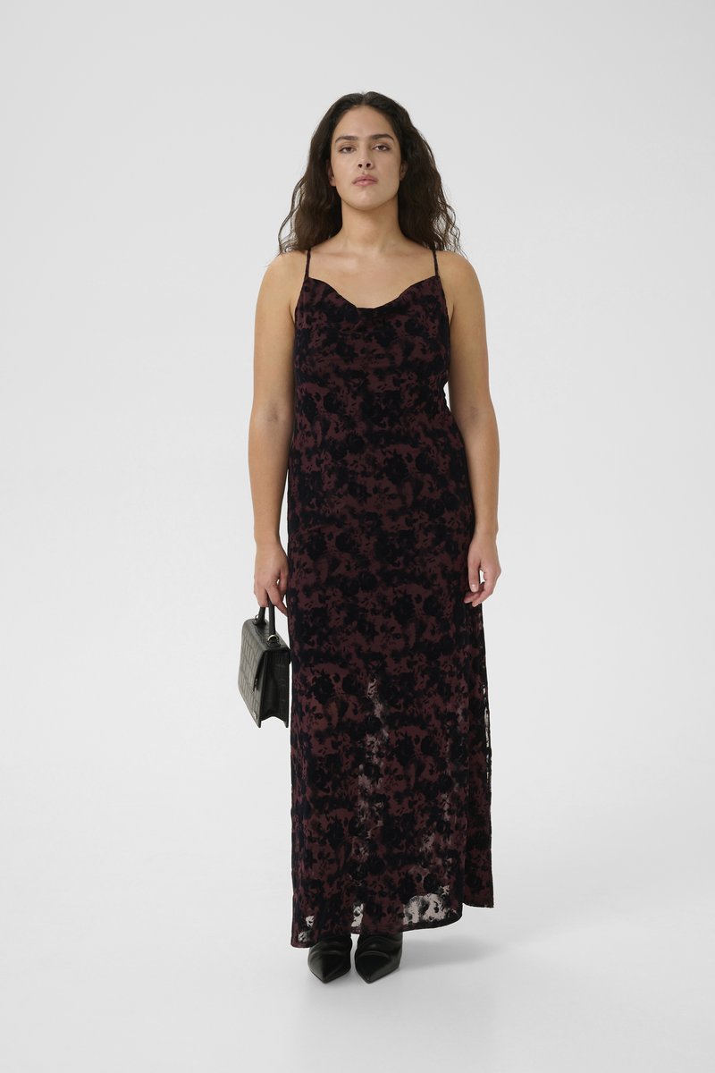 Maxi dress with thin straps, featuring a dark floral pattern in shades of burgundy and black. Model holding a small black handbag, wearing pointed shoes.