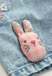 Punto di coniglio a quadretti rosa con accenti floreali, cucito su tessuto denim blu con una finitura testurizzata, caratterizzato da piccoli fiori ricamati.