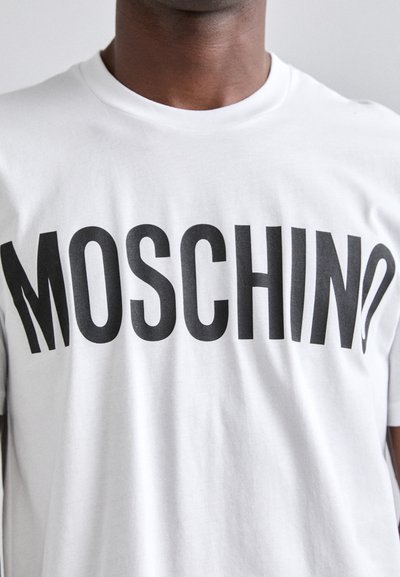 Balta medvilnės marškinėliai su ryškiu juodu "MOSCHINO" užrašu priekyje. Apvalus iškirpimas, trumpomis rankovėmis ir laisvas siluetas.