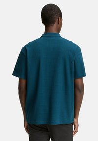 Rövid ujjú teal színű pólóing, textúrázott anyagból készült. Klasszikus gallérral és lapos alsó szegéllyel rendelkezik; a hátoldalon nincs látható márkajelzés.