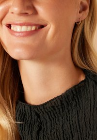 Gouden oorring met gestructureerd detail, gedragen door een persoon met lang blond haar. Zwarte, gerimpelde stof bij de hals is gedeeltelijk zichtbaar.
