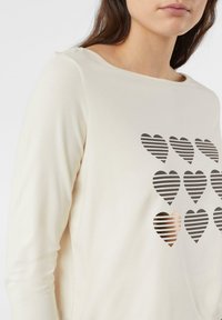 Kremowa bluzka z długim rękawem, zdobiona wzorem czarnych i metalicznych paskowanych serc na przodzie. Miękki materiał z okrągłym dekoltem.