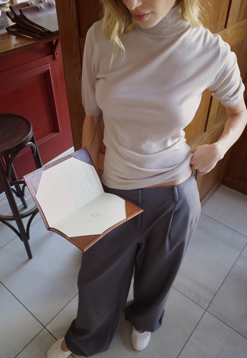 Topa corta beige claro, pantalones anchos gris oscuro, sosteniendo un menú de cuero marrón con páginas blancas. Taburete de madera y pared texturizada de fondo.