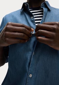 Camicia in denim con design a bottoni, colore azzurro chiaro e tessuto leggermente strutturato. Sotto, è visibile una maglietta a righe in bianco e nero.