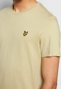 Beżowy bawełniany t-shirt z okrągłym dekoltem, krótkimi rękawami i żółto-czarnym haftowanym logo motyla na lewej piersi.