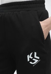 Μαύροι φόρμες με ελαστική μέση και λευκό λογότυπο KLJ Karl Lagerfeld Jeans κοντά στην τσέπη, όπου ένα χέρι είναι μερικώς εισαχθεί.