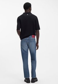 Tricou polo negru din tricot cu mâneci scurte, asortat cu blugi din denim albastru deschis, având o etichetă roșie cu logo pe spate.