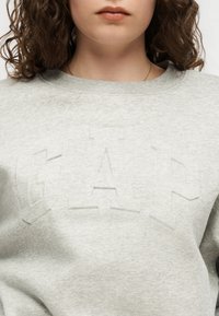 Άτομο που φοράει ανοιχτό γκρι φούτερ με διακριτικό ανάγλυφο λογότυπο "GAP", ορατό από τους ώμους έως λίγο κάτω από το πηγούνι, με κατσαρά μαλλιά.