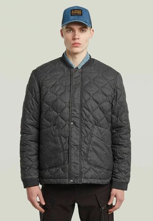 Junger Mann trägt eine schwarze gesteppte Jacke, ein blaues Jeanshemd und eine blaue Mütze mit Logo und steht vor einem schlichten hellen Hintergrund.