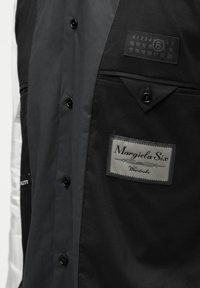 Černá vícepanelová košile s knoflíkovým zapínáním a kapsami. Obsahuje látkovou etiketu, potištěné grafiky a hladkou texturu. Stříbrné detaily na rukávu.