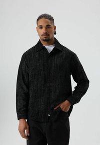 Zwart corduroy overhemd met een kraag, knoopsluiting aan de voorkant en lange mouwen. Gestructureerd oppervlak en een ontspannen pasvorm, gedragen over een wit shirt.