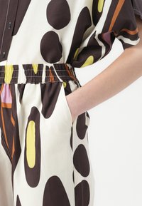Lässiger Jumpsuit mit einem braunen, cremefarbenen und bunten abstrakten Muster. Elastischer Taillenbund, kurze Ärmel und seitliche Taschen.