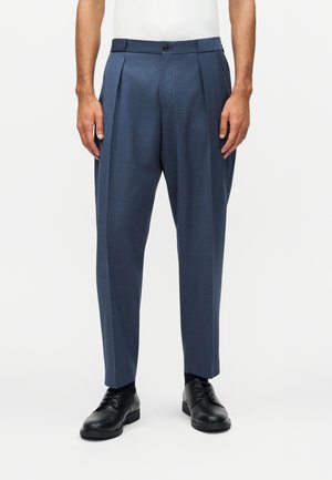 Άνδρας φορεί μπλε ραμμένο παντελόνι με πιέτες και μαύρα δερμάτινα επαγγελματικά παπούτσια, στέκεται μπροστά σε απλό λευκό φόντο.