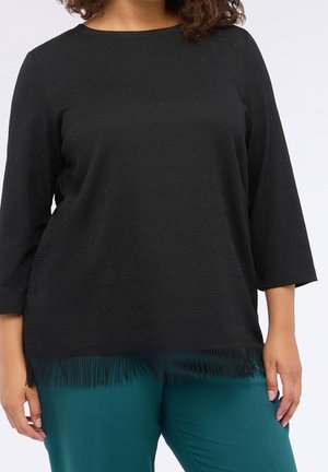 Kvinde iført en sort langærmet top med frynsekant og turkise bukser, vist fra halsen til hoften mod en hvid baggrund.
