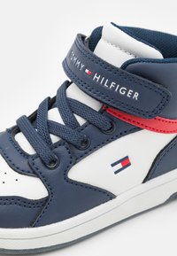 Zapatilla alta en navy y blanca con una correa de Velcro, que presenta acentos rojos y azules, cuero texturizado y un logotipo en la correa y el costado.