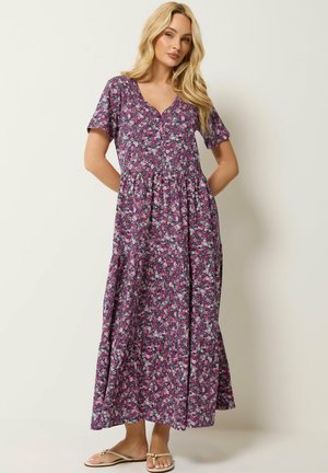 Femme blonde portant une robe longue violet à fleurs à manches courtes et des sandales beige, debout devant un fond clair, les mains dans le dos.