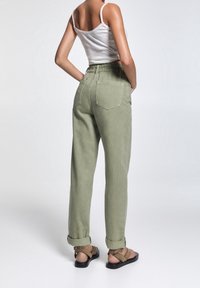 Mujer que lleva pantalones de cintura alta verde oliva con los puños remangados, una camiseta sin mangas blanca y sandalias planas marrones, de pie con las manos en los bolsillos.