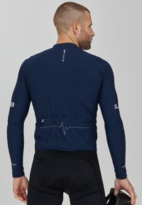 Marineblaues Langarm-Radtrikot aus elastischem Material mit pulsierenden Linien auf dem Rücken und reflektierenden Akzenten an den Ärmeln, kombiniert mit schwarzen Shorts.